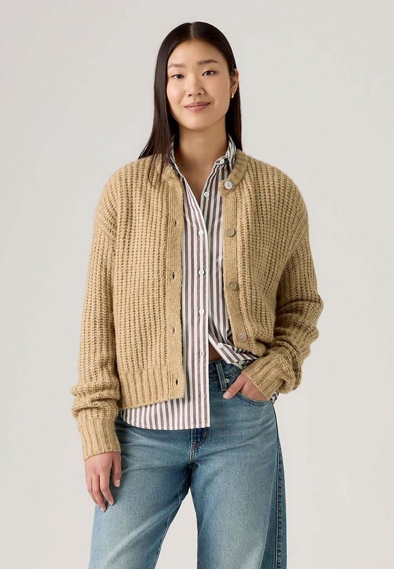 Cardigan a maglia beige con un motivo testurizzato, scollo rotondo e chiusura con bottoni; abbinato a una camicia bianca a righe con bottoni e jeans.