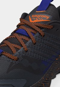 Κοντινή λήψη μαύρου παπουτσιού τρεξίματος Saucony με κορδόνια μαύρα με πορτοκαλί στίγματα και μπλε λεπτομέρειες στη γλώσσα και στη σόλα.