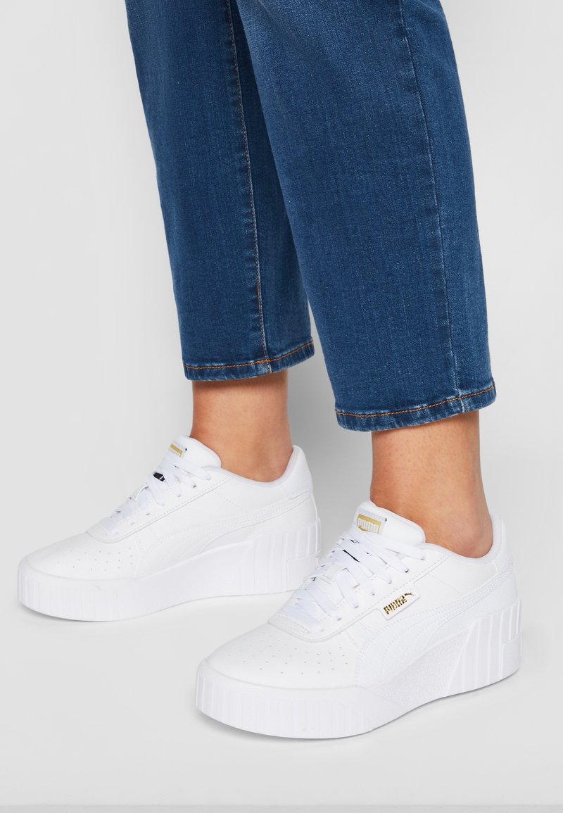 Personne portant des baskets blanches Puma à plateforme associées à un jean court bleu, sur un fond gris clair.
