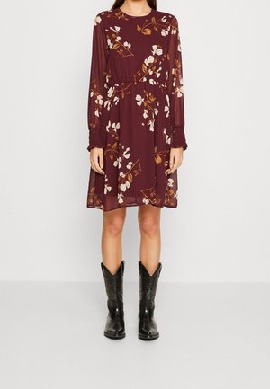 Femme portant une robe longue à manches longues à fleurs couleur bordeaux, associée à des bottes de cowboy en cuir noir, debout contre un mur blanc uni.