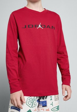 Barn iført rød langærmet skjorte med sort "JORDAN" tekst og hvid Jumpman-logo, kombineret med hvide bukser med flerfarvede Jordan-logoer.
