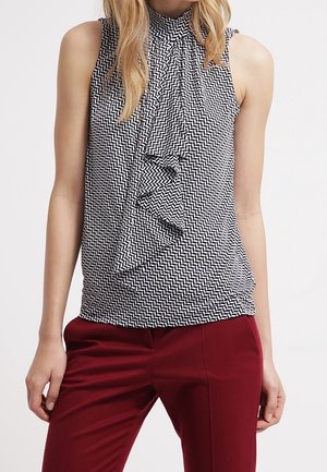 Blouse sans manches à motifs noir et blanc avec des détails volantés, portée avec un pantalon taille haute rouge foncé.