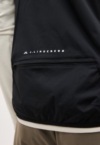 Μαύρο σακάκι με φερμουάρ σε τσέπη και λευκό λογότυπο "J.LINDEBERG" πάνω από την τσέπη, φορεμένο πάνω από ρουχισμό σε μπεζ και καφέ χρώματα.