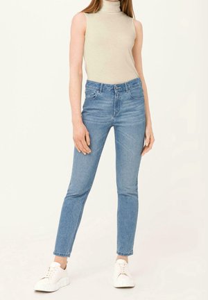 Kvinde iført beige ærmeløs rullekravetop, lyseblå højtaljede skinny jeans og hvide sneakers, står mod en ensfarvet baggrund.