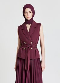 Gilet sans manches bordeaux avec des boutons dorés à double boutonnage et une ceinture à nouer, associé à une jupe plissée bordeaux. Le tissu a une texture lisse.