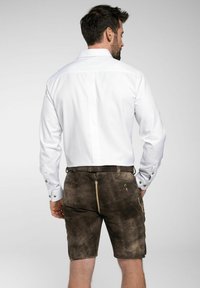 Weißes Hemd mit klassischem Kragen, kombiniert mit braunen Wildleder-Shorts, die über Seitentaschen und einen sichtbaren Reißverschluss verfügen.