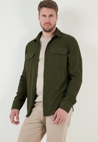 Cămașă de corduroy verde închis cu două buzunare pe piept, închidere cu butoane, mâneci lungi, țesătură texturată, purtată peste un tricou bej și pantaloni bej.