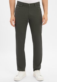 Man wearing dark green straight-leg pants and white sneakers standing against a plain white background.