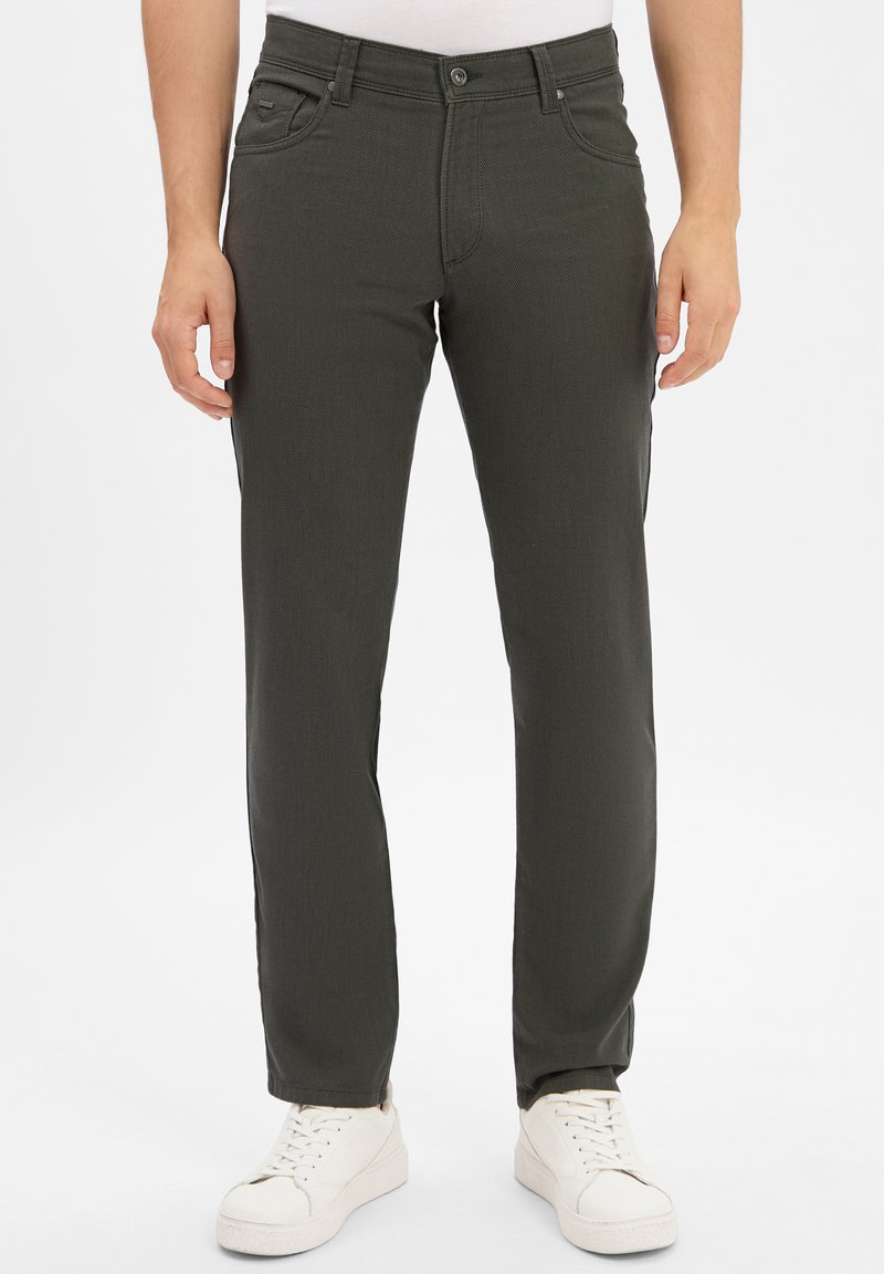 Man wearing dark green straight-leg pants and white sneakers standing against a plain white background.