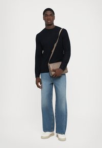 Mann kledd i en svart strukturert genser, lyseblå vid jeans, hvite sneakers og holder en beige crossover-veske foran en hvit bakgrunn.