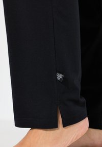 Pantalons en coton noirs avec une fente sur le côté à la cheville. Présente une petite étiquette de marque tissée près de l'ourlet. Texture lisse.