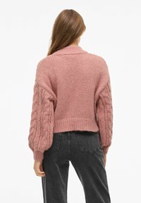 Chunkasta roza pletena majica z rokavi v kabelskem pletenju in rebrastim robom. Nosi se čez črne hlače z visokim pasom, ki imajo sproščen kroj.