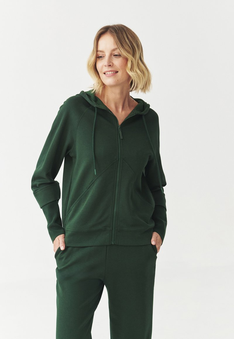 Sudadera verde con cremallera, hecha de un tejido suave, con capucha, dos bolsillos frontales y puños acanalados. Combinada con pantalones verdes a juego.