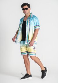 Satin-Hemd mit kurzen Ärmeln in einem blauen bis gelben Farbverlauf, Regenbogenstreifen, kombiniert mit gelben Shorts mit ähnlichen Streifen und Logo. Schwarze Sneaker.