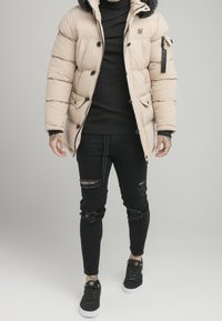 Beige Pufferjacke mit einem mit Fell gefütterten Kapuze, schwarzen Akzenten, mehreren Knöpfen und Reißverschlusstaschen. Getragen über einem gerippten schwarzen Pullover und verwaschenen schwarzen Jeans.