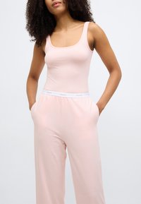 Világos rózsaszín ujjatlan felső mély nyakkivágással, laza rózsaszín nadrággal párosítva, amelynek fehér derékpántján "HUGO" logó található. Sima szövettextúra.