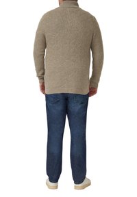 Beiger Strickpullover mit strukturiertem Muster, hohem Kragen und langen Ärmeln, kombiniert mit blauen Jeans und hellen Sneakers.