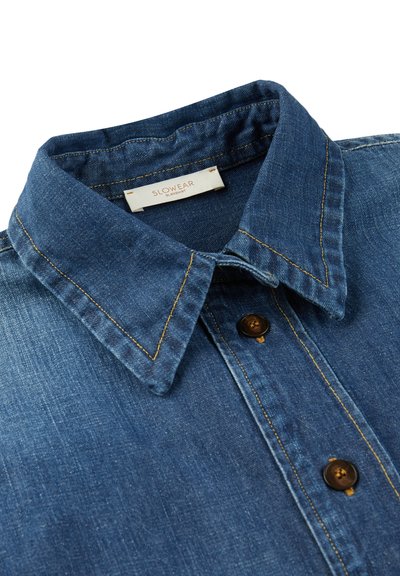 Denim overhemd met een gestructureerde kraag, donkerblauwe stof, contrasterende gele stiksel, en bruine knopen, met een label dat "SLOWEAR" zegt.