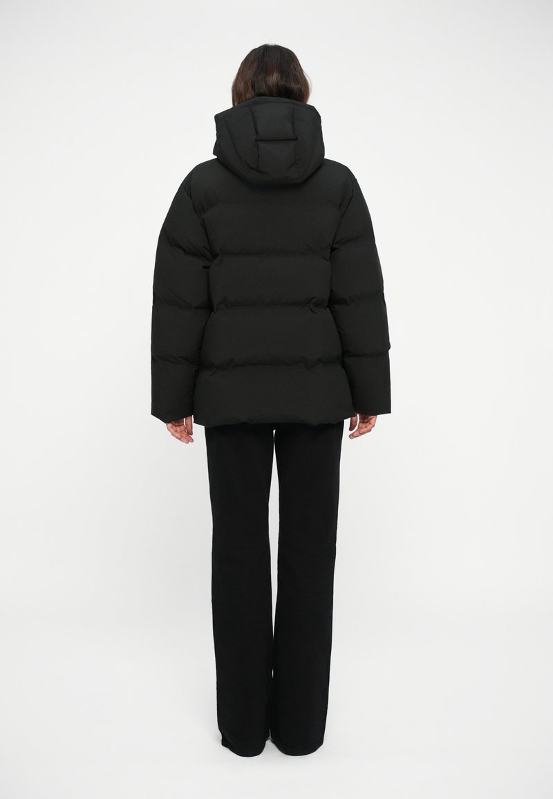 Doudoune noire avec de grandes sections matelassées et une capuche. Le dos a une coupe droite, complétée par un pantalon noir ajusté.