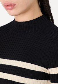 Črn rebrast pleten top z visokim ovratnikom, ki ima dve vodoravni smetano belii črtu. Tkanina se zdi mehka in ima pristen kroj.