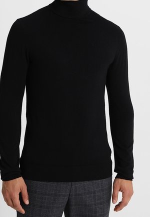 Mann trägt einen enganliegenden schwarzen Rollkragenpullover mit langen Ärmeln und graue karierte Hosen, steht vor einem schlichten Hintergrund.