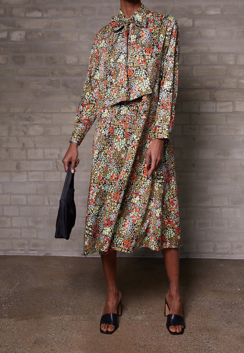 Robe midi fleurie à manches longues, col noué et jupe évasée dans des tons de vert, rouge et noir, portée avec des sandales plates noires.