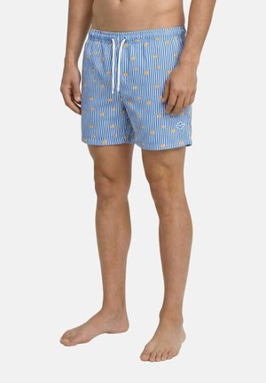 Mand iført blå og hvide vertikalt stripede shorts med små orange frugt mønstre og hvid snor, stående barfodet.