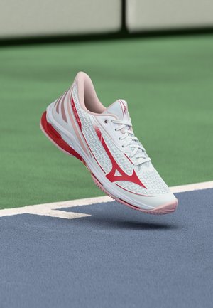 White and red athletic shoe hovering above a tennis court with green and blue surface and white boundary lines.