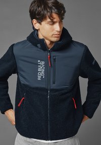 Marineblauwe jas van fleece-stof, met een ritssluiting aan de voorkant, een capuchon en rode ritsstoppers. Bevat een logo en twee zijzakken. Zachte textuur.