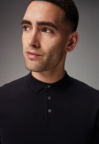Camisa polo negra con cuello, tres botones y una tela texturizada, mostrando un primer plano de la parte superior del torso y la cara de un modelo masculino.