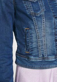 Jeansjacke mit tailliertem Design, verblasster blauer Farbe und sichtbaren Nähten. Verfügt über eine Seitentasche und strukturierte Stoffe.