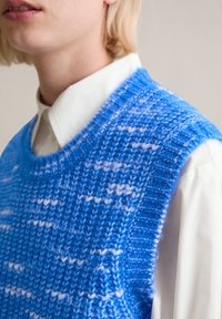 Niebieski sweter w formie kamizelki, dzianinowy, o teksturowanym wzorze, z okrągłym dekoltem i ściągaczami na brzegach, noszony na białej koszuli z kołnierzykiem.