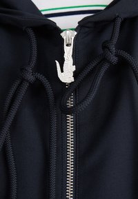 Jūrų laivyno zip-up džemperis su sidabro spalvos krokodilo tipo užtrauktuku, kontrastingais raišteliais ir lygiu audinio tekstūra.