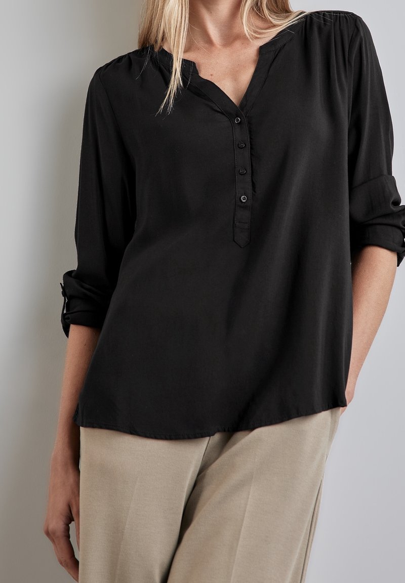 Femme portant un chemisier noir à manches longues boutonné avec les manches retroussées et un pantalon beige, debout devant un fond neutre.