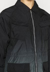 Giacca nera con zip frontale, due tasche sul petto e un tessuto testurizzato con un effetto ombre che va da tonalità scure a tonalità più chiare.