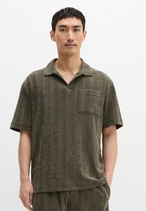 Jonge man draagt een donkerolijfgroen, gestructureerd polo met korte mouwen en een borstzakje, staand tegen een effen witte achtergrond.