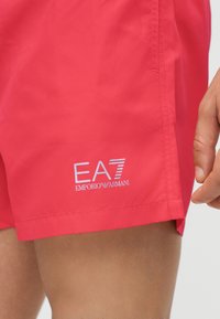 Pantaloni scurți roșii aprinși cu logo alb EA7 Emporio Armani pe coapsa stângă, purtați de o persoană cu mâna și partea superioară a piciorului vizibile.
