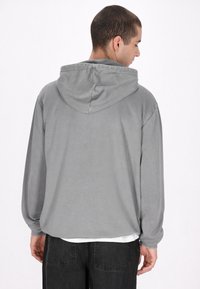 Szürke, hosszúujjú kapucnis pulóver, laza szabással, húzózsinóros kapucnival, bordás mandzsettákkal és puha felülettel. Fehér alsó póló felett viselve.