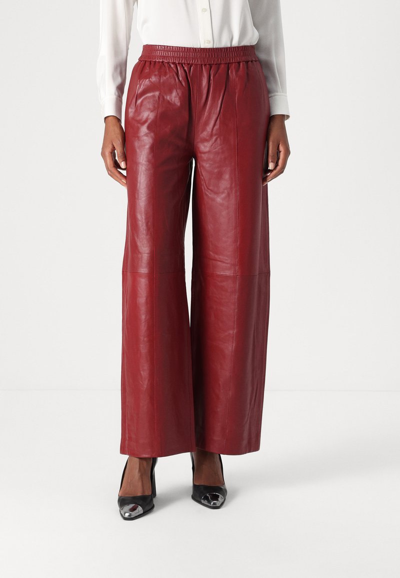 Pantalons larges en cuir rouge avec une taille élastique, texture lisse et détails de couture visibles. Associés à des chaussures à talons hauts noires.