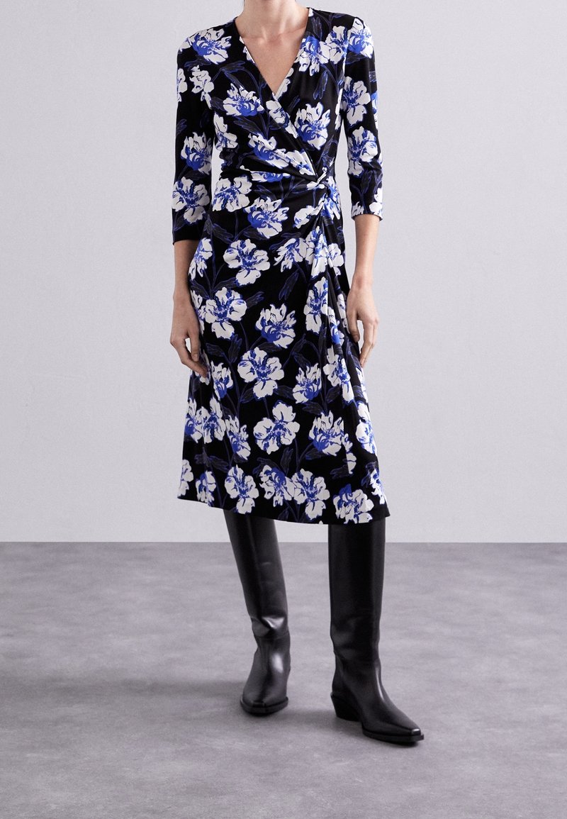 Femme portant une robe portefeuille noire avec des motifs floraux bleus et blancs et des manches trois-quarts, associée à des bottes noires mi-mollet.