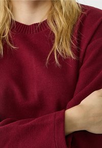 Blonde persoon in een kastanjebruine gebreide trui met geribde kraag, met de arm over de borst gevouwen, close-up op het torso- en nekgebied.