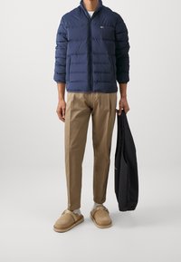 Gewatteerd marineblauw jack met ritssluiting, beige broek en bruine instapschoenen. Houdt een zwarte tas vast, staat tegen een lichtgrijze achtergrond.