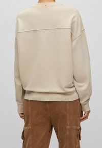 Beige vä oversized-collegepaita, jossa värillinen korostus takana, resorit hihansuissa ja helmassa, yhdistettynä ruskeisiin puuvillaisiin cargo-housuihin.