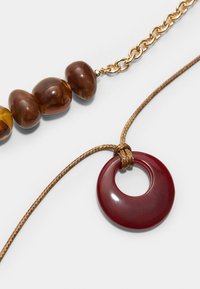 Collana con perle marroni lucide e un pendente circolare color bordeaux, infilata su un cordino intrecciato marrone con un accento di catena dorata.