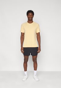 Ανοιχτό κίτρινο κοντομάνικο μπλουζάκι, μαύρο σορτς και λευκά αθλητικά παπούτσια. Το ύφασμα φαίνεται λείο· απλός σχεδιασμός χωρίς ορατά σχέδια ή λογότυπα.