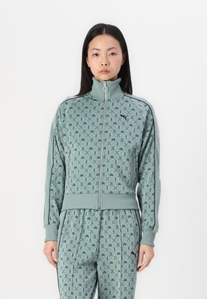 Veste de survêtement zippée verte avec un col haut, présentant un motif géométrique et des poignets côtelés, associée à un pantalon assorti à motif.