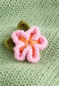 Háčkovaná růžovo-oranžová hvězdicovitá květina se dvěma zelenými listy ležící na texturované světle zelené pletené tkanině.