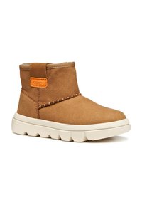 Botines de ante marrón con una suela de goma blanca, detalles de costura y una etiqueta naranja en el lateral. Textura suave con punta redondeada.