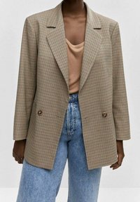 Blazer marrón de cuadros con solapas grandes, doble botonadura y un corte relajado, combinado con un top beige y vaqueros azules de talle alto.