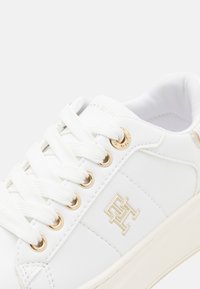 Sneaker bianco con un esterno in pelle liscia, dettagli dorati sui passanti e un logo impresso sul lato. Presenta lacci bianchi e un interno imbottito.